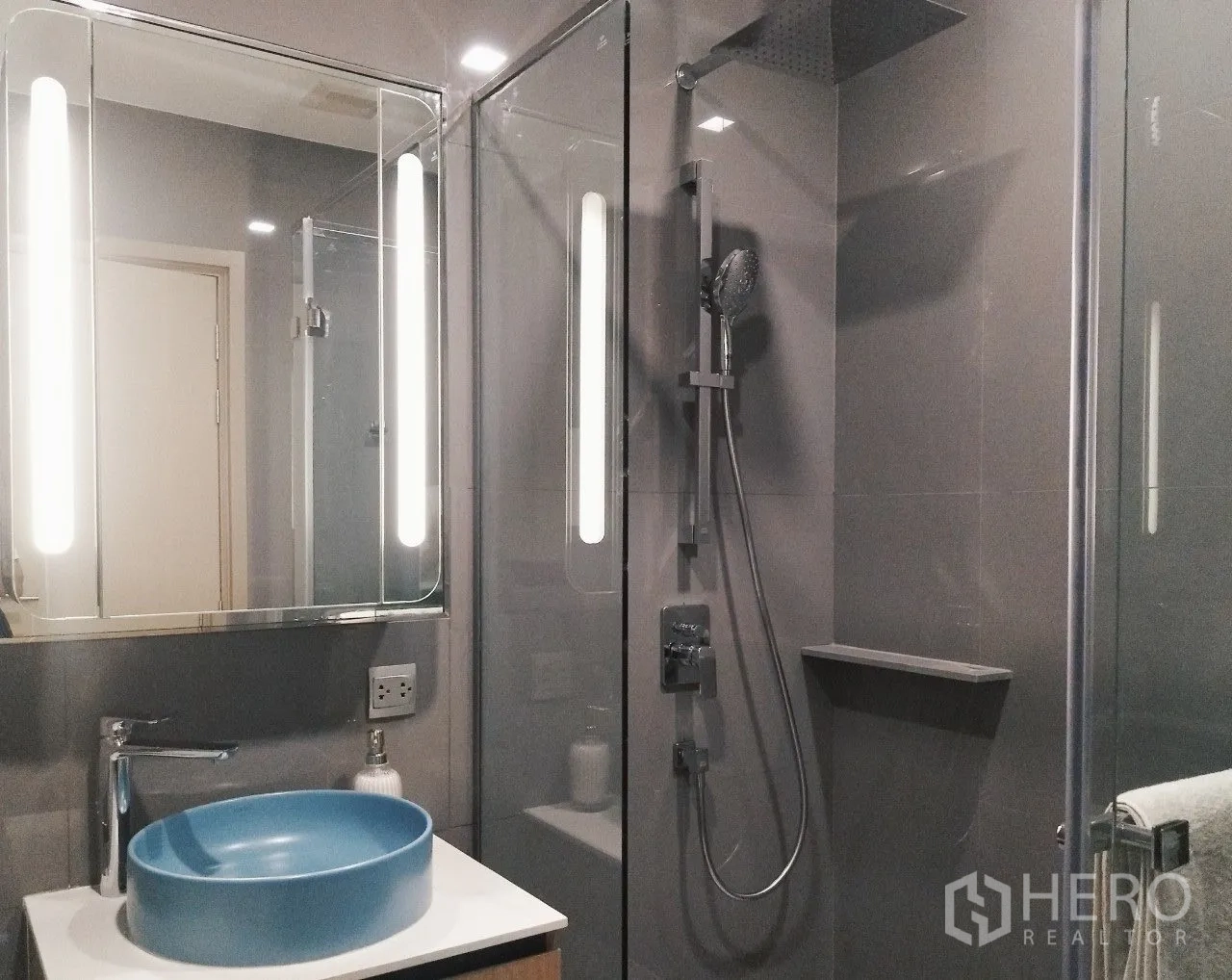 Condominium for rent in Watthana, Bangkok - Glass-enclosed shower with rain head beside a lit mirror and blue vessel sink.