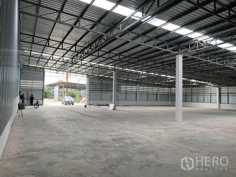 Warehouse for rent in Mueang Pathum Thani, Pathum Thani - Clear-span warehouse interior with steel truss roof, skylight panels and concrete floor, wide drive-in gate at the rear.