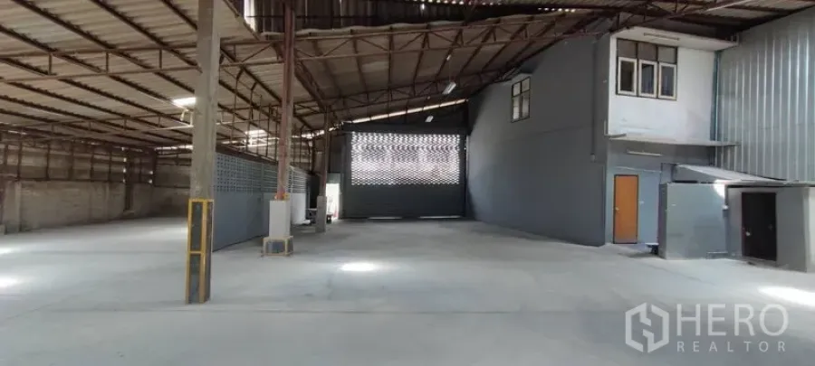 Warehouse for rent in Phra Khanong, Bangkok - Open-plan warehouse interior with high steel truss roof, concrete floor, and an internal mezzanine office beside a large loading gate.