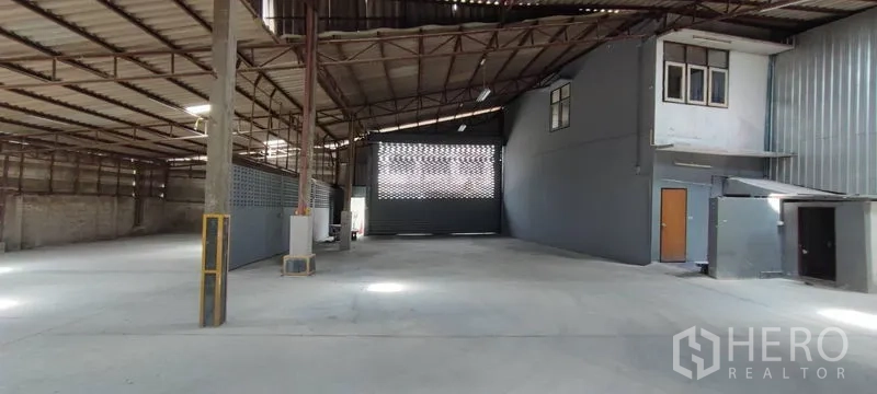 Warehouse for rent in Phra Khanong, Bangkok - Open-plan warehouse interior with high steel truss roof, concrete floor, and an internal mezzanine office beside a large loading gate.