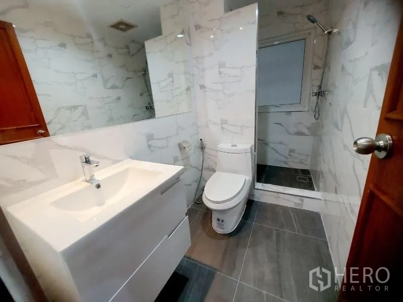 Condominium for rent in Watthana, Bangkok - Compact bathroom with vanity, toilet and walk-in shower featuring marble walls and gray floor tiles.
