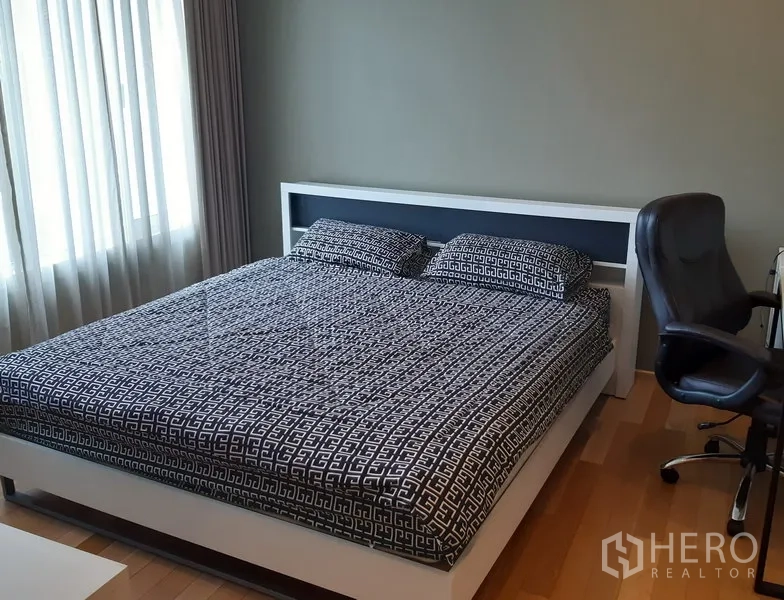 Condominium for rent in Khlong Toei, Bangkok - Bedroom with large bed, work desk and natural light through curtained window on wooden floors.