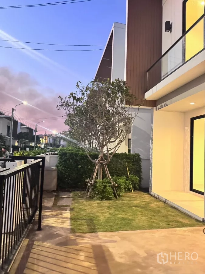 Detached House for rent in Samut Prakan - Front garden with sprinkler, walkway and neighboring modern homes at sunset.
