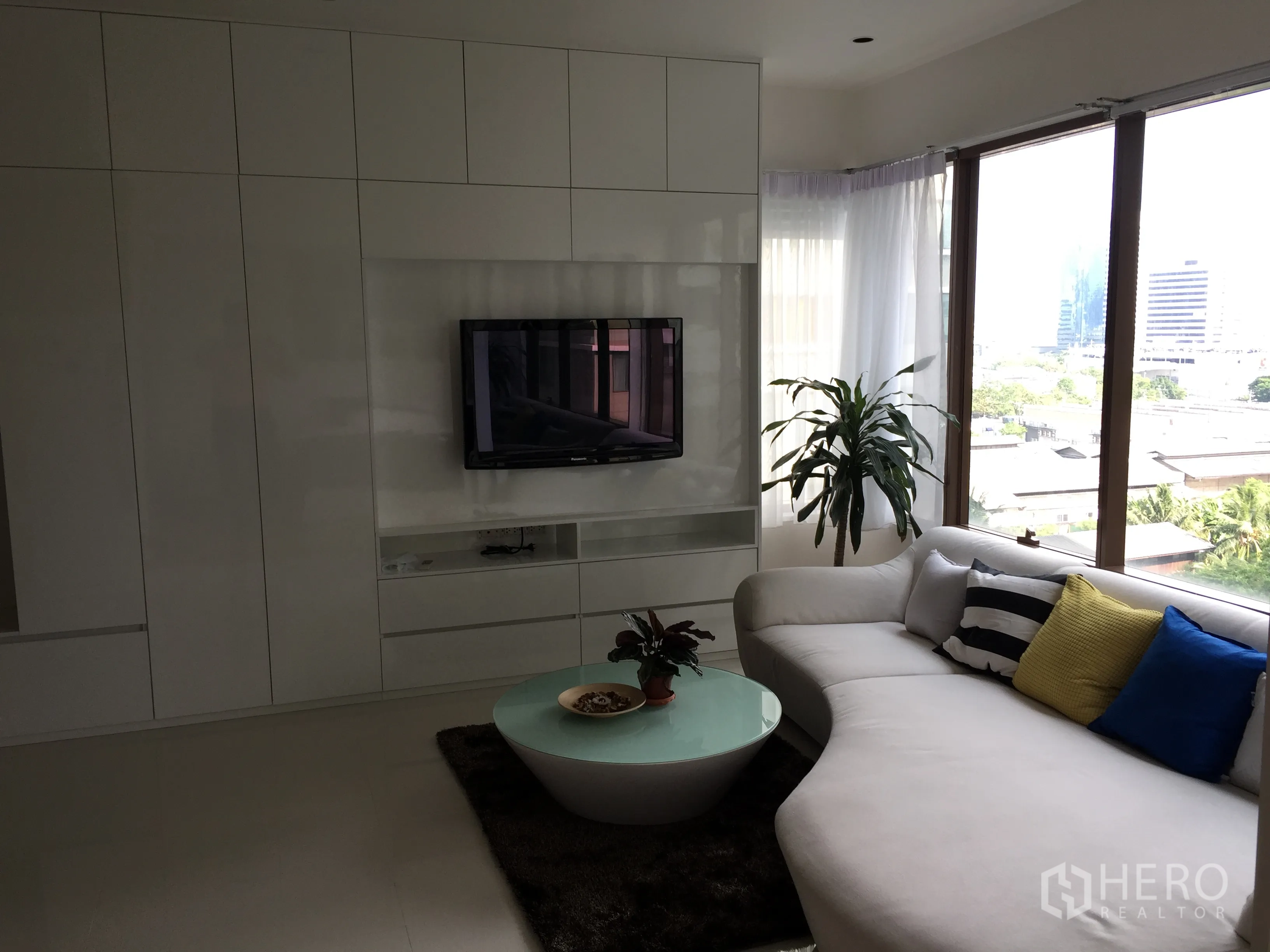 Condominium for rent in Khlong Toei, Bangkok - Living room with built‑in TV wall, sectional sofa, indoor plant and panoramic windows overlooking the city.