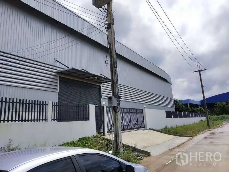 Factory for rent in Samut Prakan - Side view of the warehouse facade with roller shutter door and security fence.