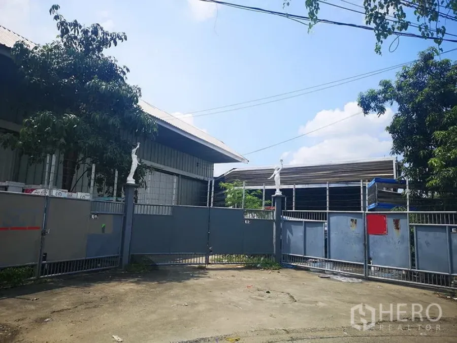 Warehouse for rent in Bang Khen, Bangkok - Secure warehouse entrance with sliding metal gate and shade trees in Bangkok.