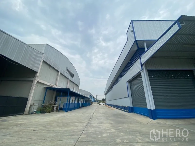 货栈 出租在Pathum Thani - 两栋现代钢结构仓库之间的宽阔混凝土通道，带装卸平台和卷帘门。