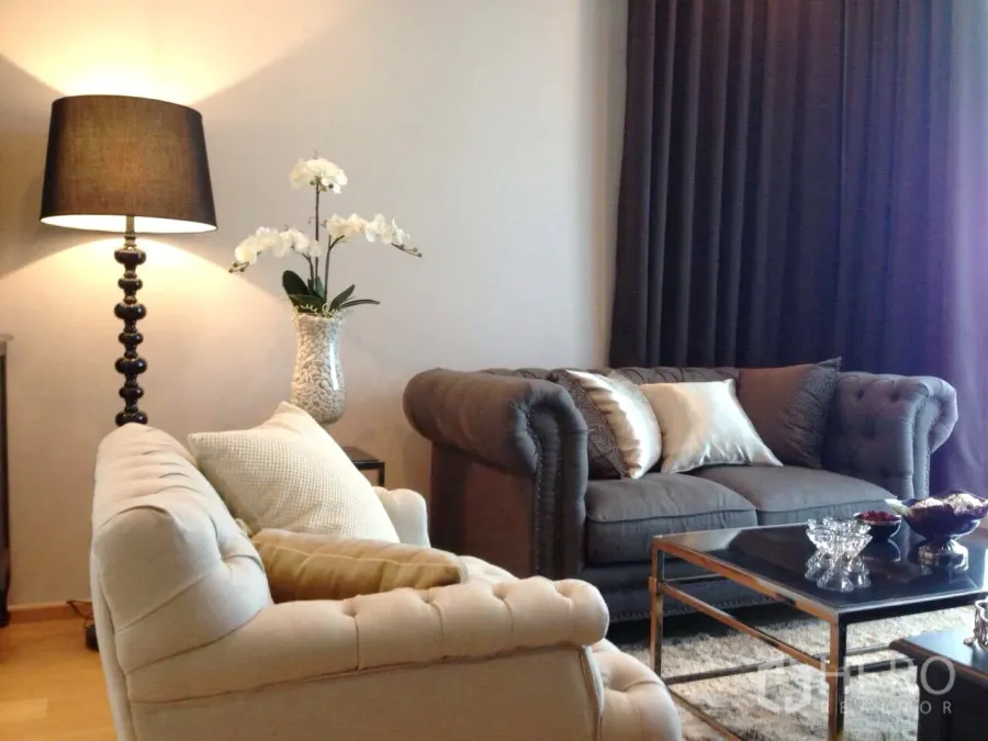 Condominium for rent in Khlong Toei, Bangkok - Elegant living room with tufted sofas, floor lamp, dark curtains and glass-top coffee table.