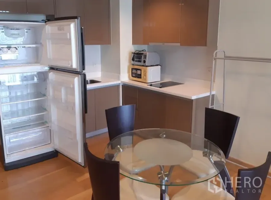 Condominium for rent in Khlong Toei, Bangkok - Modern kitchenette with brown cabinets, open refrigerator and a round glass dining table.