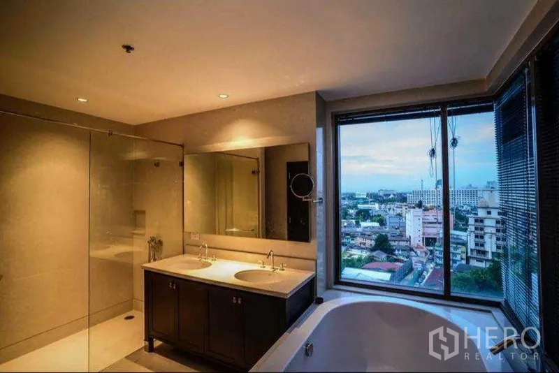 Condominium for rent or sale in Khlong Toei, Bangkok - Luxury bathroom with double vanity, separate glass shower and bathtub overlooking the city skyline.