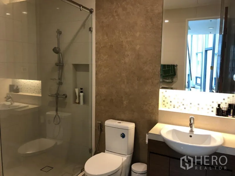 Condominium for rent or sale in Khlong Toei, Bangkok - Modern bathroom with glass-enclosed shower and mosaic-lit mirror.