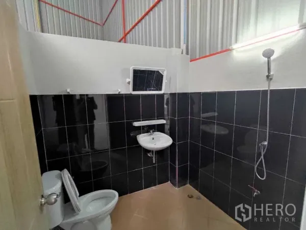Warehouse for rent in Pak Kret, Nonthaburi - Warehouse restroom with toilet, wall-mounted sink, and shower against black tiled walls.