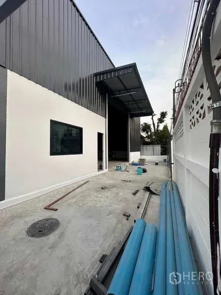 Warehouse for rent in Bang Khen, Bangkok - Side yard of the warehouse with grey facade, awning, and utility pipes on a wide concrete walkway.