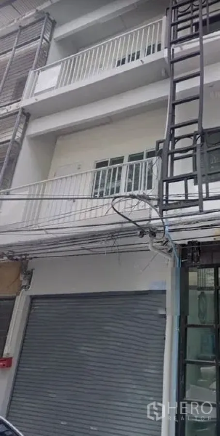 Shophouse for rent in Khlong Toei, Bangkok - Four-storey shophouse facade with roller shutter shopfront, metal-railed balconies, grey-white exterior and visible street wiring.