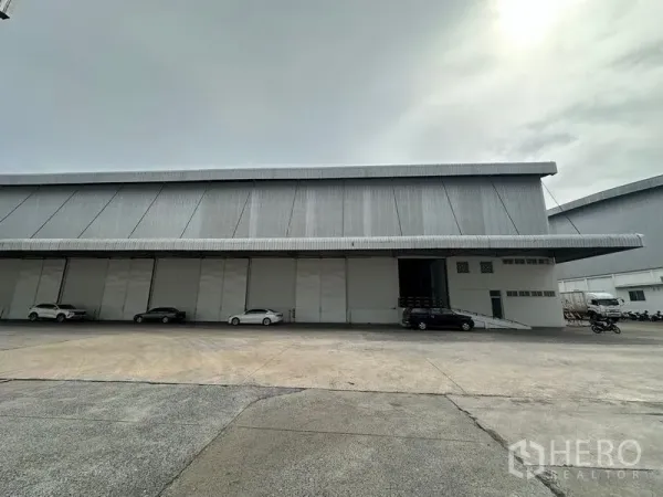 Warehouse for rent in Phra Nakhon Si Ayutthaya - Large metal-clad warehouse exterior with long loading bay, ramp and parked vehicles.