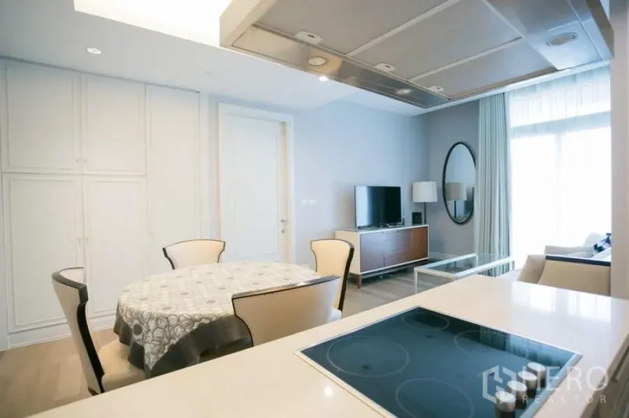Condominium for rent or sale in Pathum Wan, Bangkok - Open-plan living area with round dining table, TV console and kitchen island cooktop beside a large window.