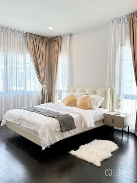 独立式住宅 出租或出售在Bang Phli, Samut Prakan - 明亮臥室，軟包床頭、床頭櫃與深色木地板上的毛絨地毯。