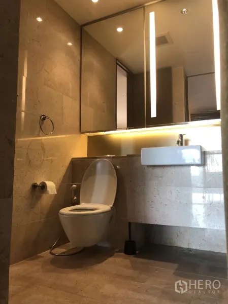 Condominium for rent in Pathum Wan, Bangkok - Modern bathroom with stone tiles, illuminated mirror, vanity sink, and contemporary toilet.