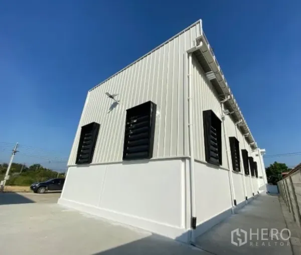 Warehouse for sale in Sai Noi, Nonthaburi - Side view of the warehouse showing black louvered ventilation windows, wall lights, and ample parking area.