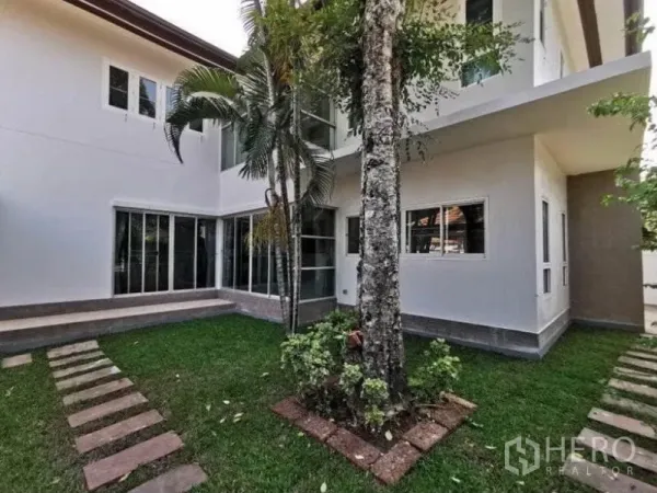 Detached House for rent in Bang Kapi, Bangkok - Green courtyard lawn with a mature tree beside a white two‑storey exterior and corner sliding doors.