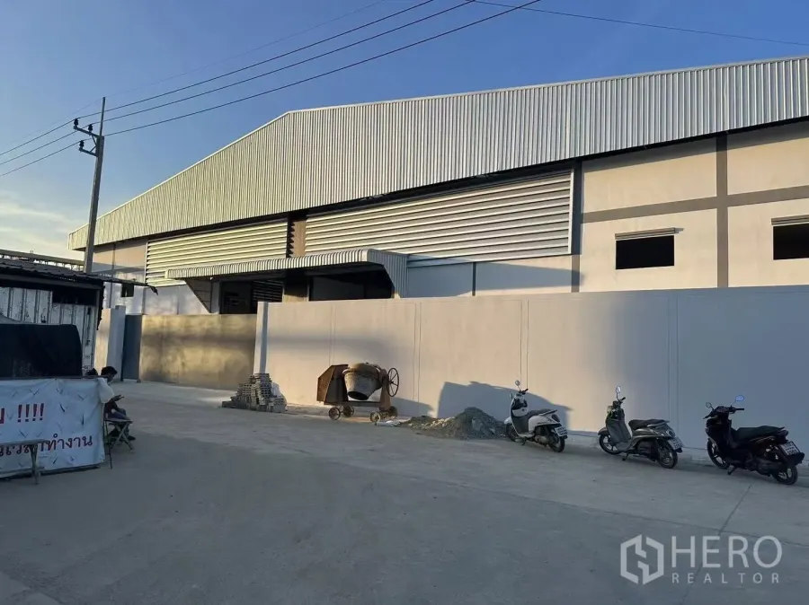 Factory for rent in Nong Khaem, Bangkok - Modern factory exterior with high metal roof, solid perimeter wall and wide concrete drive at sunset.