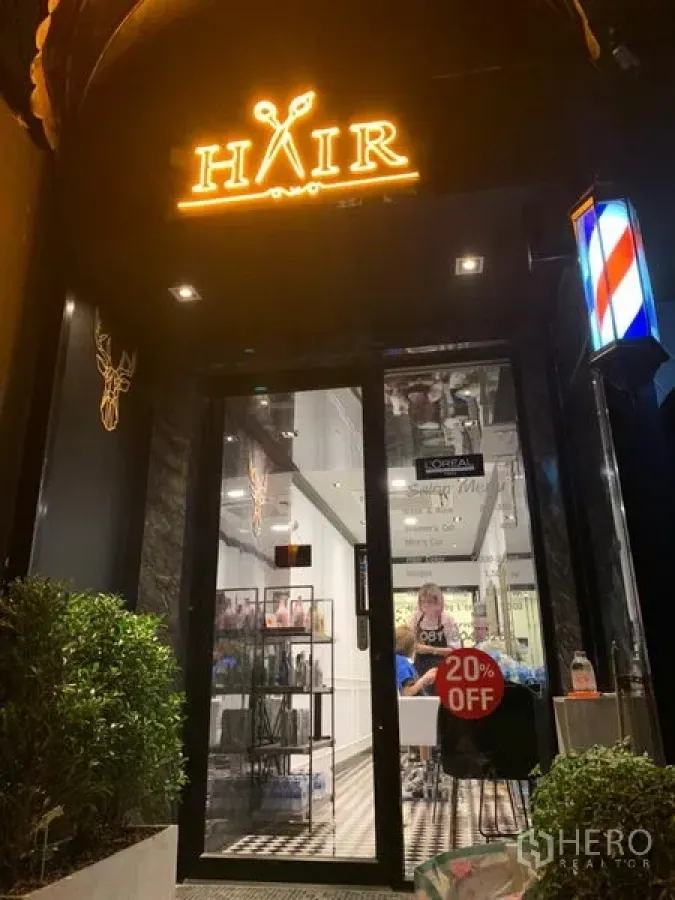 Retail Space for rent in Watthana, Bangkok - Night view of a retail shop entrance with an illuminated HAIR sign, glass door, barber pole, and greenery.