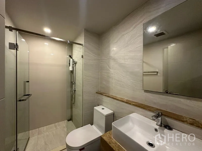 Condominium for rent in Khlong Toei, Bangkok - Contemporary bathroom featuring glass-enclosed shower, vanity with basin, and toilet.