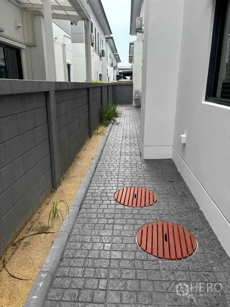 Detached House for rent in Mueang Nonthaburi, Nonthaburi - Side walkway with stamped concrete path, boundary wall and utility access covers.