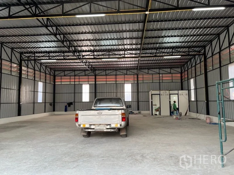 Warehouse for rent in Bueng Kum, Bangkok - Open 300 sqm warehouse interior with high steel truss roof, concrete floor and bright strip lighting, with a pickup truck parked inside.
