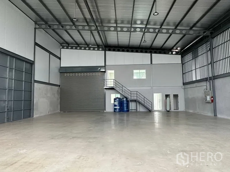 Warehouse for rent in Samut Prakan - Open storage area with high roof, roller shutter door, small windows and utility corner.