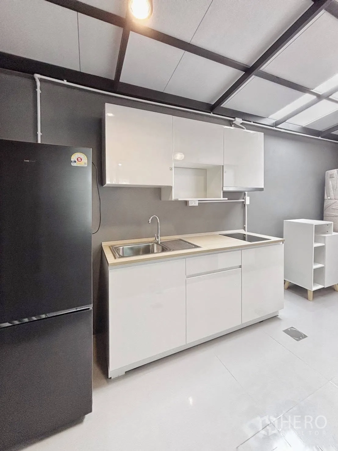 Townhouse for rent in Bang Phli, Samut Prakan - Compact fitted kitchen with white cabinets, sink, cooktop and a black refrigerator under exposed beams.