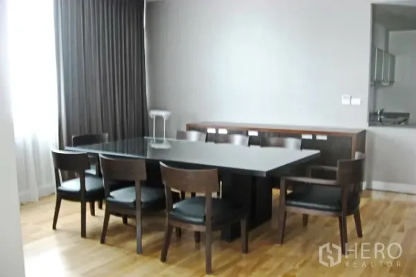 Condominium for sale in Khlong Toei, Bangkok - Eight-seat dining room with a dark table, sideboard, and floor-to-ceiling curtains next to a semi-open kitchen.