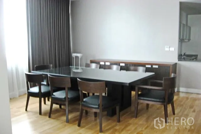 Condominium for sale in Khlong Toei, Bangkok - Eight-seat dining room with a dark table, sideboard, and floor-to-ceiling curtains next to a semi-open kitchen.