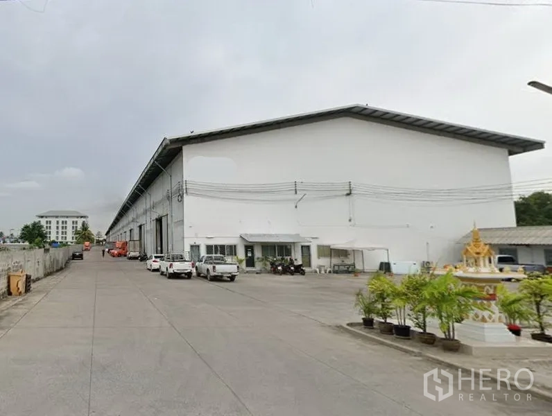 Warehouse for rent in Prawet, Bangkok - Large white warehouse exterior in Prawet, Bangkok with wide concrete driveway, multiple loading bays, parking and potted greenery.