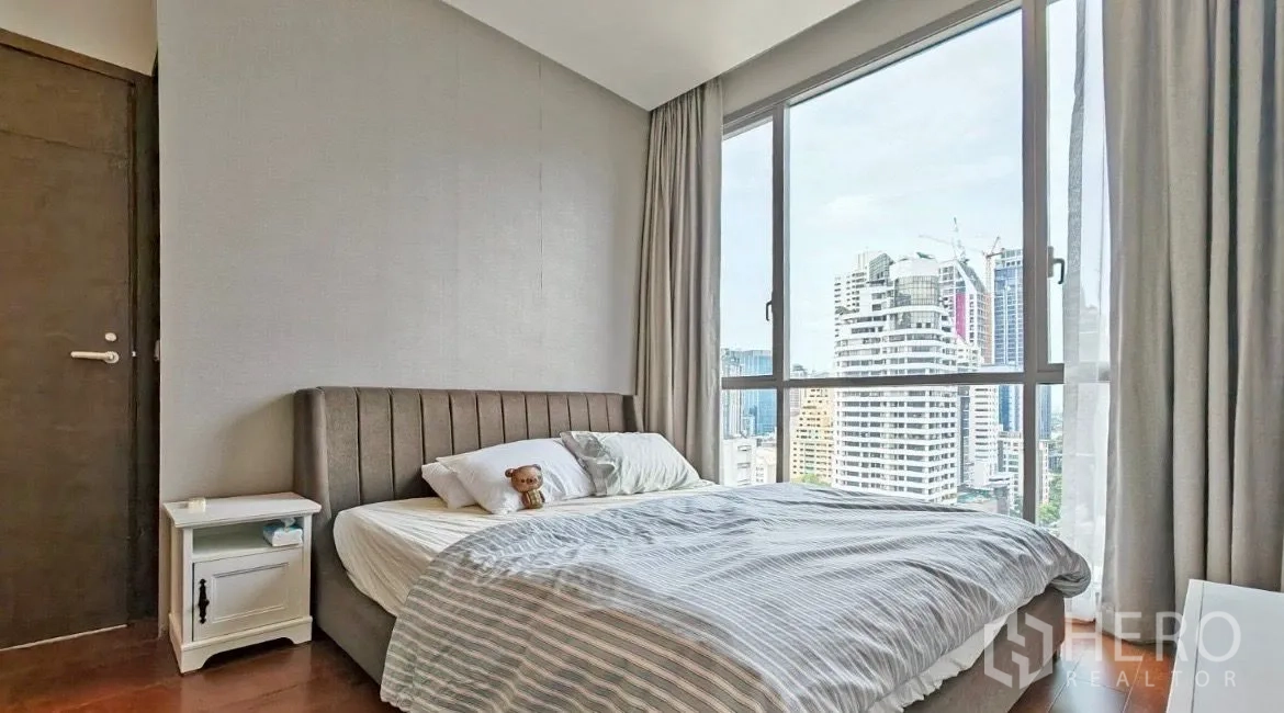 Condominium for rent in Watthana, Bangkok - Bedroom with upholstered headboard and bedside table, featuring a large window and panoramic skyline outlook.