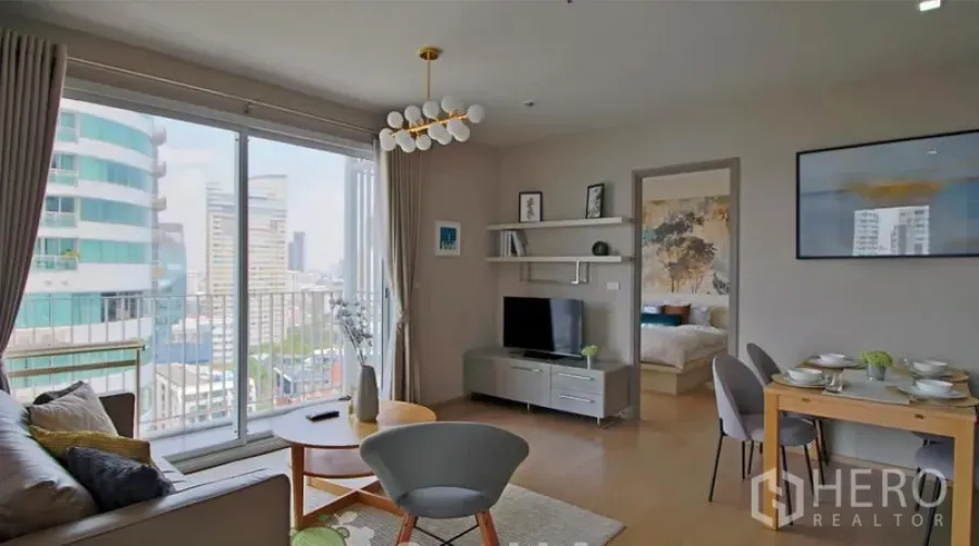 Condominium for rent in Watthana, Bangkok - Living room with city-view balcony, TV console, dining table, and a doorway leading to the bedroom.