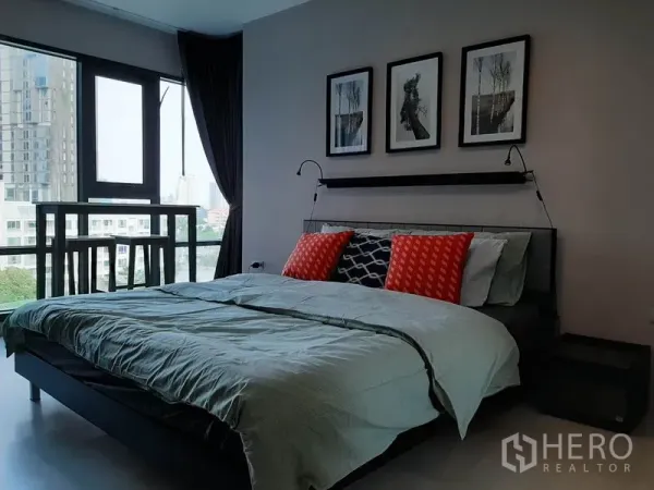 Condominium for rent or sale in Khlong Toei, Bangkok - Bedroom featuring a city view through large windows and a neatly made bed with colorful pillows.