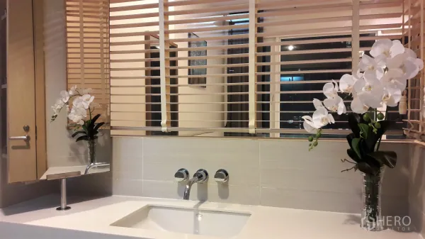 Condominium for rent in Khlong Toei, Bangkok - Modern bathroom vanity with chrome fixtures and white orchids beside blinds that open to the bedroom.