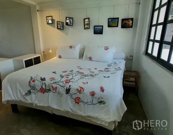 Townhouse for rent in Khlong Toei, Bangkok - Bright bedroom with king bed, floral duvet and hanging photo frames by corner windows.