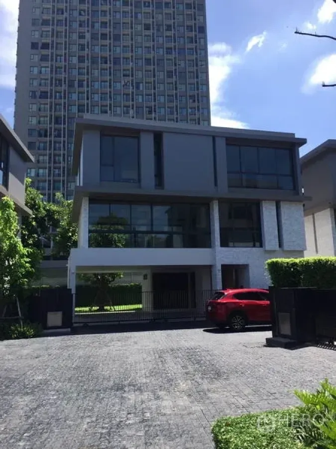 Detached House for rent in Watthana, Bangkok - Contemporary three-storey detached house with floor-to-ceiling windows, gated driveway and a red car, with a high-rise tower behind.