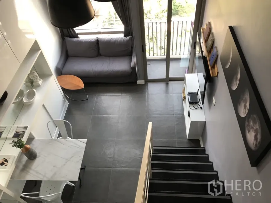 Condominium for rent or sale in Khlong Toei, Bangkok - Double-height living room with gray sofa, balcony doors, TV console, marble dining table and staircase on dark tiled floors.