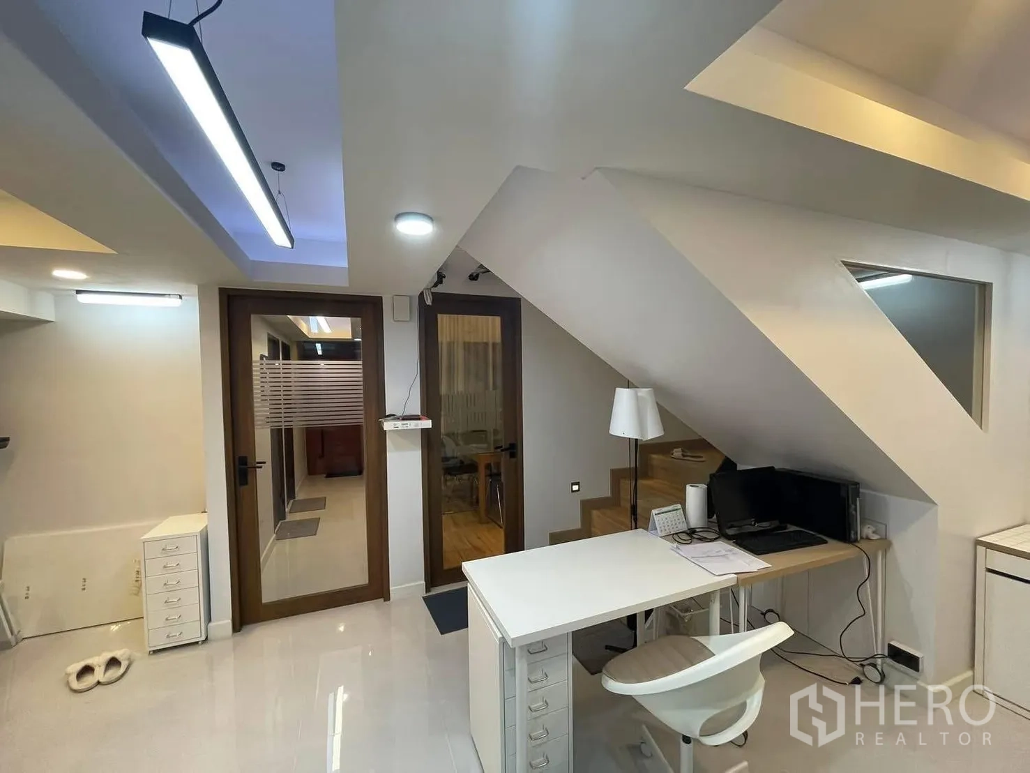 Home Office for rent or sale in Bang Kapi, Bangkok - Ground-floor workspace with built-in desk under the stairs, glass doors and modern ceiling lights.