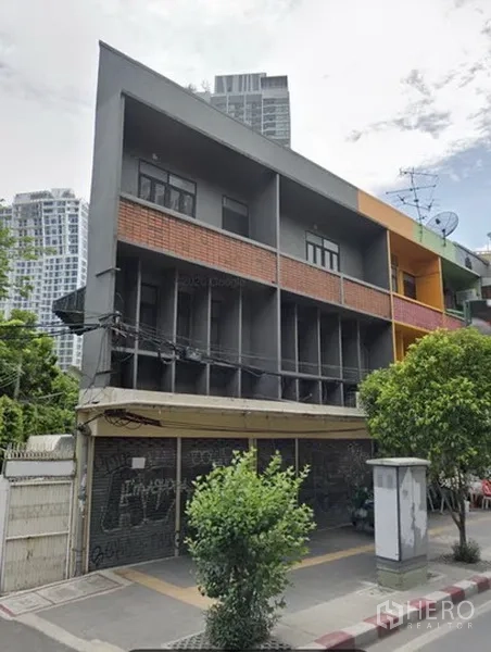 Shophouse for rent in Khlong Toei, Bangkok - Modern multi-storey shophouse with grey and brick facade on a corner plot, ground-floor roller shutters, street trees, and nearby high-rise condos in Ekkamai, Bangkok.