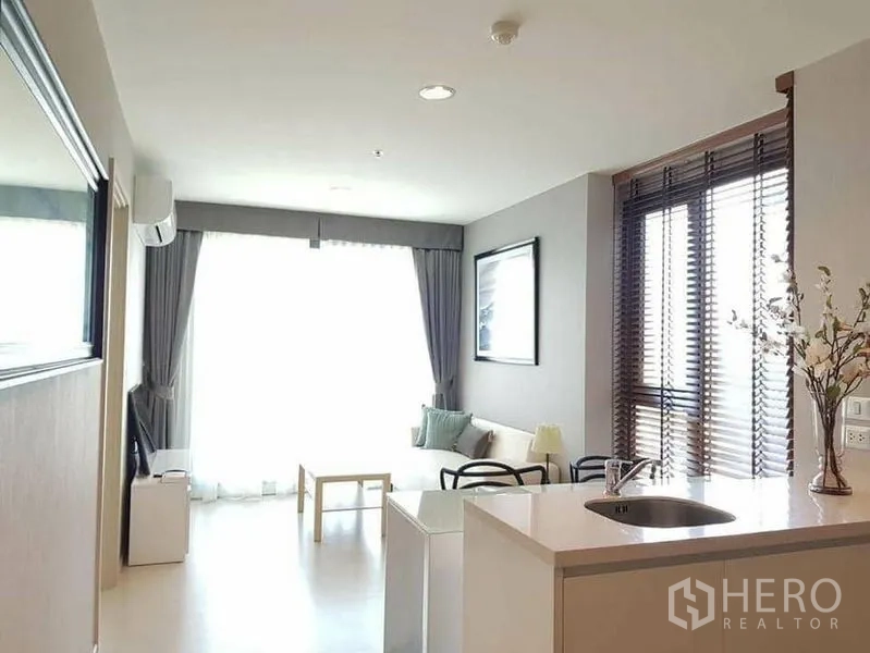 Condominium for rent or sale in Khlong Toei, Bangkok - Bright open-plan living room with kitchen island, sofa and large window with curtains.