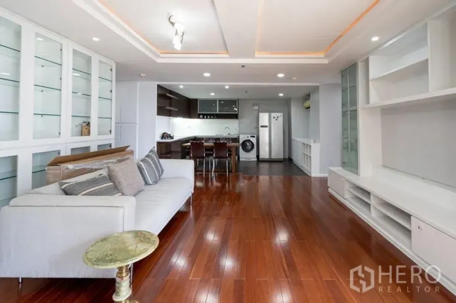 Condominium for sale in Watthana, Bangkok - Spacious open‑plan living and kitchen zone with glass display shelving and warm wood floors.