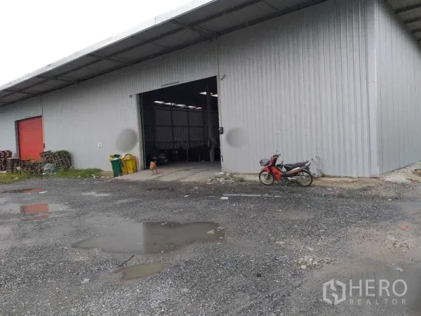 Warehouse for rent in Pak Kret, Nonthaburi - Front of a steel warehouse showing a large loading bay and wide driveway with drainage puddles.