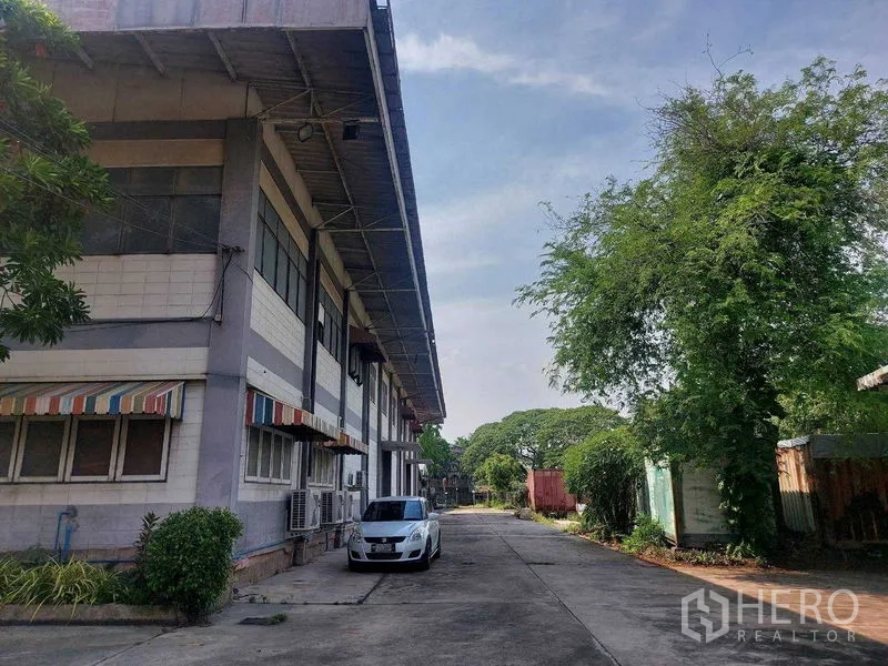 货栈 出租在Min Buri, Bangkok - 仓库侧立面，彩篷窗与车道停车位，周边绿树成荫