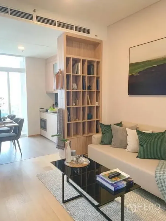 Condominium for sale in Khlong Toei, Bangkok - Bright living area with beige sofa, glass coffee table and a wooden shelving divider opening to a compact kitchen and dining space.