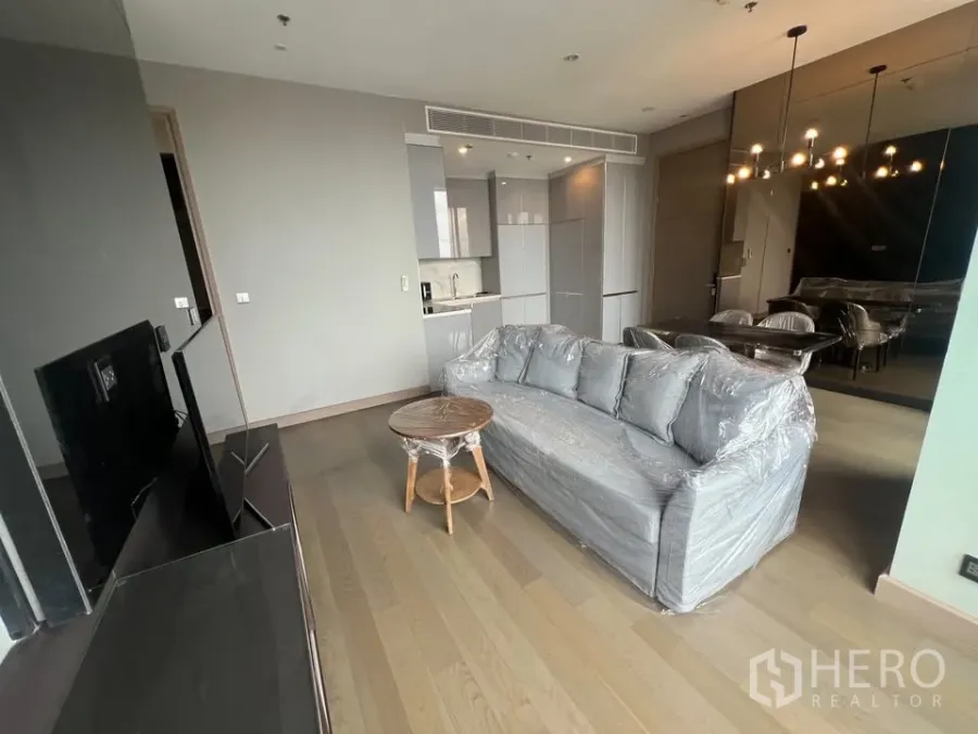 Condominium for rent in Huai Khwang, Bangkok - Open-plan living area with grey sofa, TV wall, pendant lights and compact kitchen on wood floors.