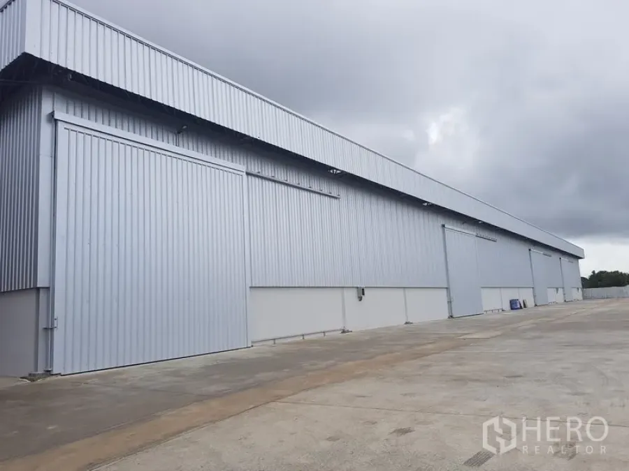 Warehouse for rent in Lat Krabang, Bangkok - Long exterior view of a metal-clad warehouse with sliding doors and a wide concrete driveway under cloudy sky.